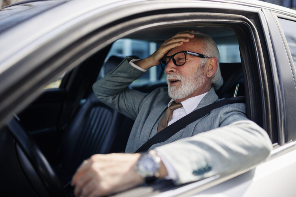 A businessman in a grey suit holds his head in frustration while driving a car.