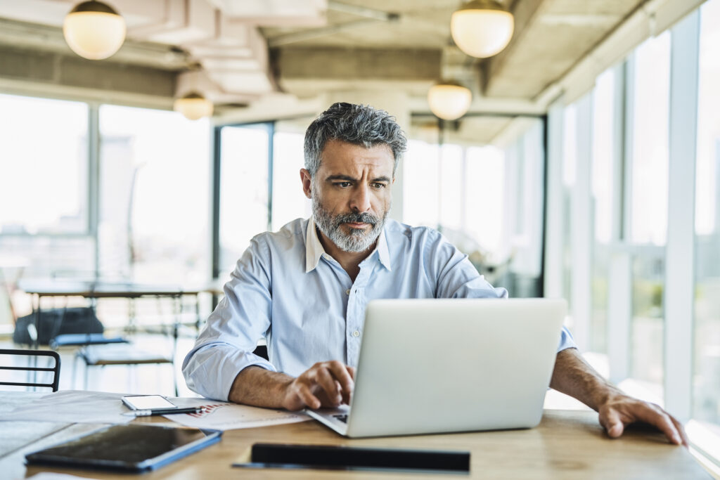 A man in a button-up shirt looks at a laptop with papers and reports around him in a sunlit coworking space.
