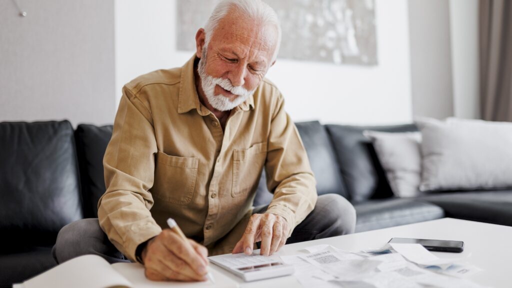 An older man smiles while he uses a calculator on his coffee table in front of his sofa.