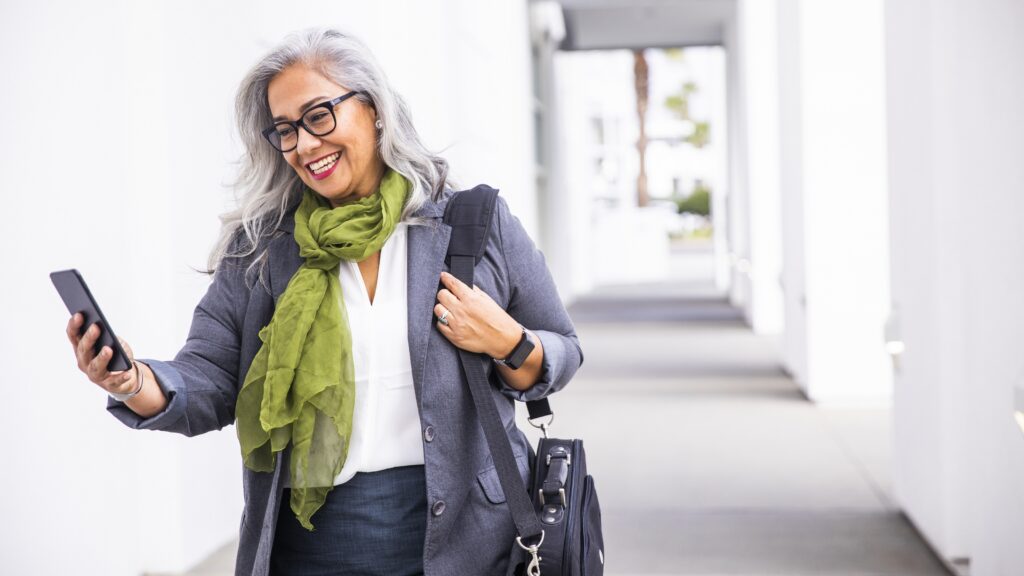 An older businesswoman smiles as she leaves the office while looking at her phone.