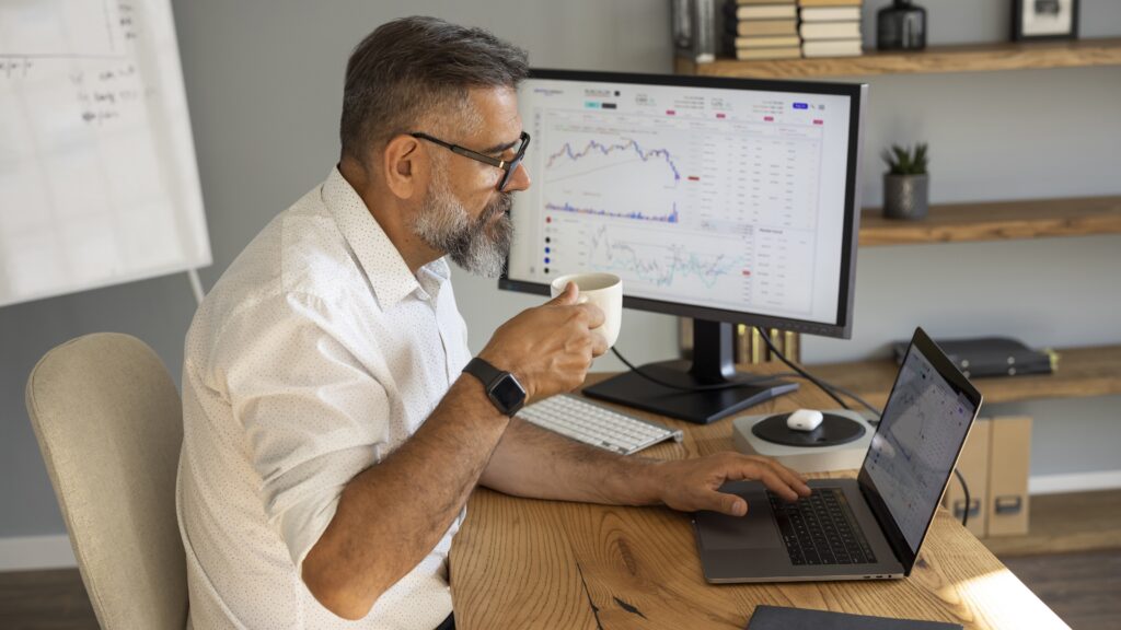 An older man looks at his laptop while sitting at his desk at home, a trading website on the monitor beside him.