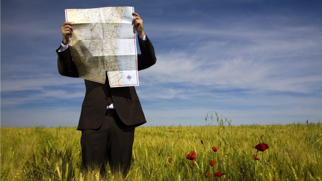 A businessman holds a map up in front of his face while standing in a field.