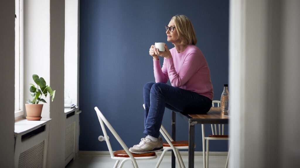 A woman sits on a table with her feet on a chair as she holds a cup of coffee and looks out the window.