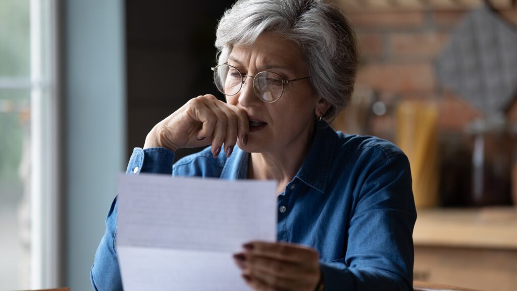 An older woman looks concerned as she looks at a letter at home.