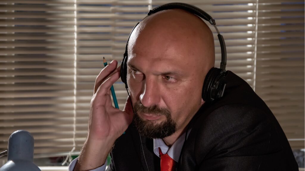 A shady-looking man wearing headphones listens in on others' conversations.