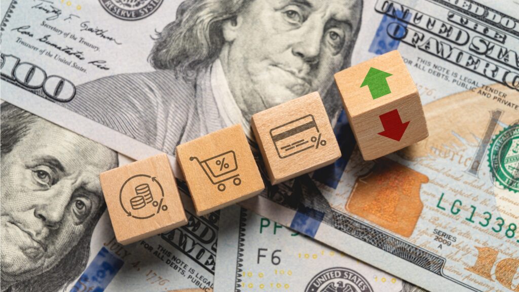 Wooden blocks representing money, shopping and a credit card sit on hundred-dollar bills next to a block with red and green arrows.