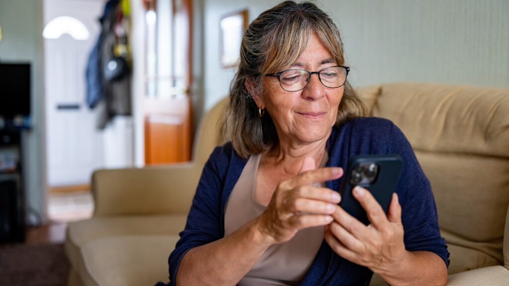 An older woman smiles as she uses an app on her phone while sitting on the sofa.