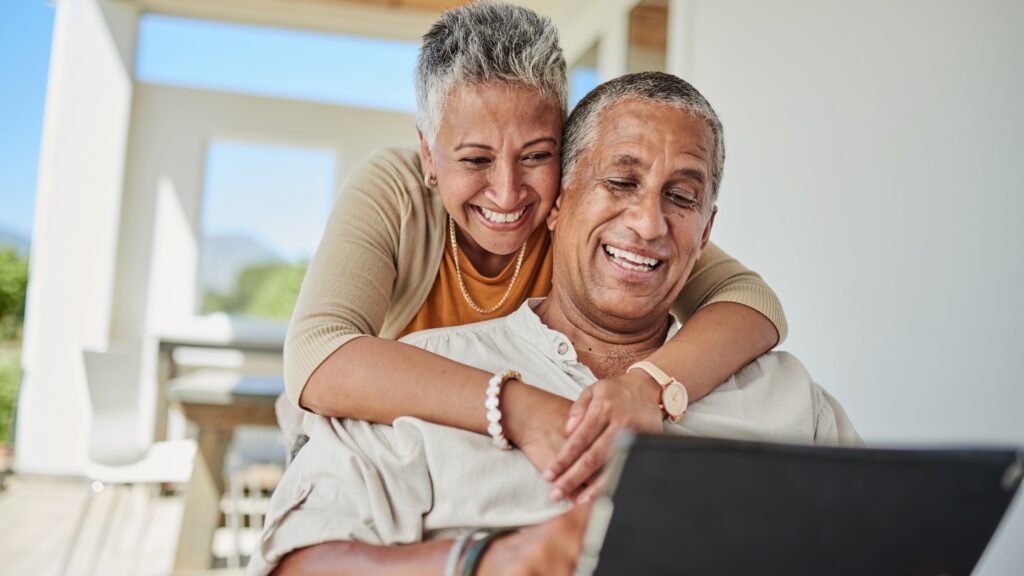 An affluent couple smile as they look at a tablet on their porch.