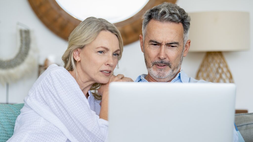 An older couple looking at a laptop together on their sofa look concerned.