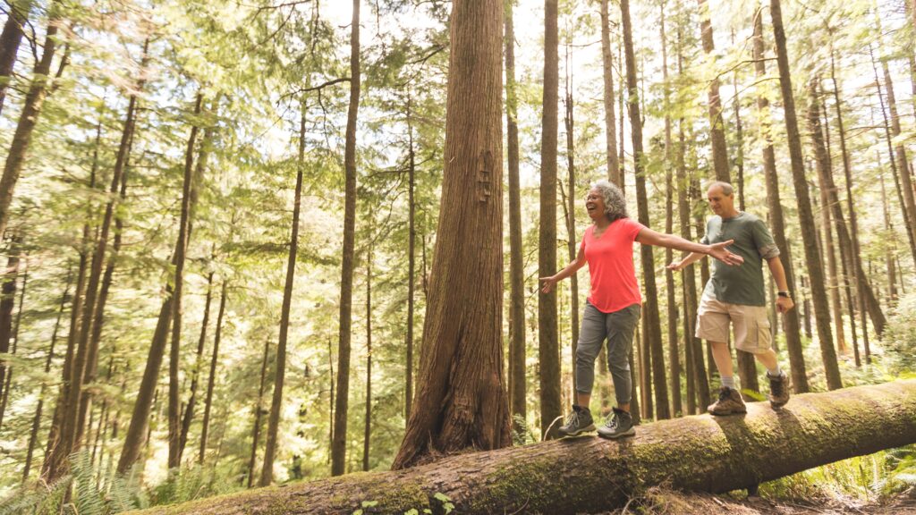 A retired couple balance on a fallen tree trunk as they hike in the woods.