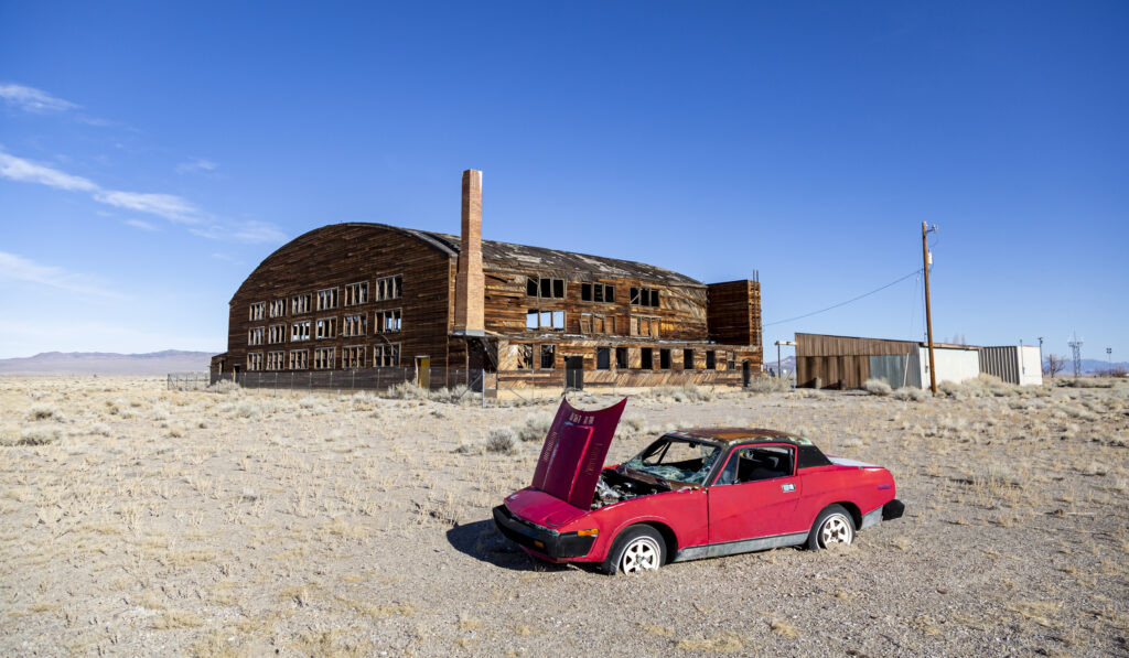 Derelict military airport hangar and retro sports car in a desert landscape, Nevada. The car's hood is up and the mood is desolate.