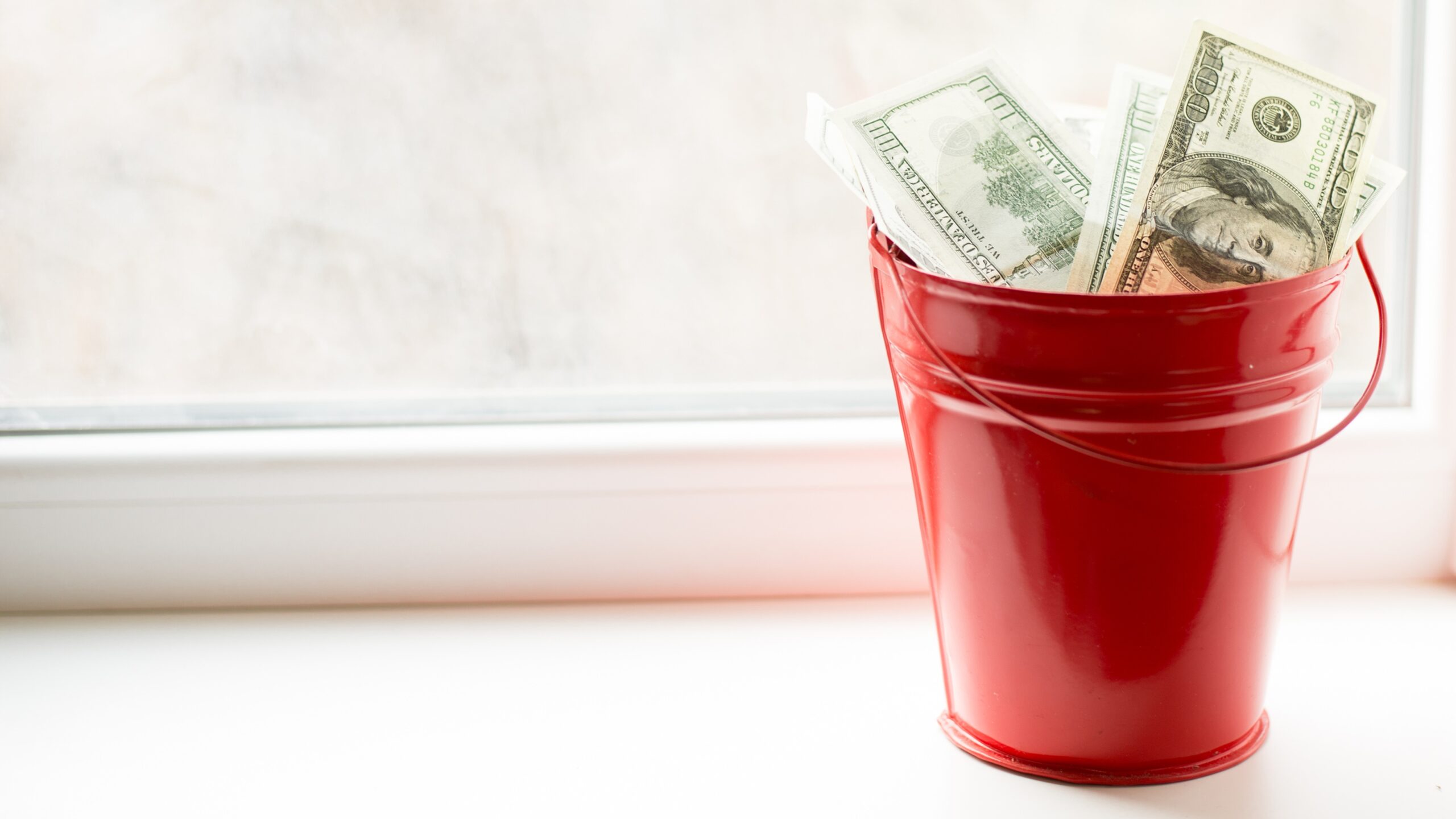 A red bucket with hundred-dollar bills in it, sitting by a window.