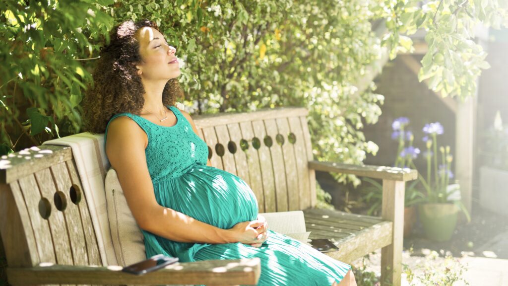 An expectant mother sits on a park bench with her head back, enjoying the sunshine.