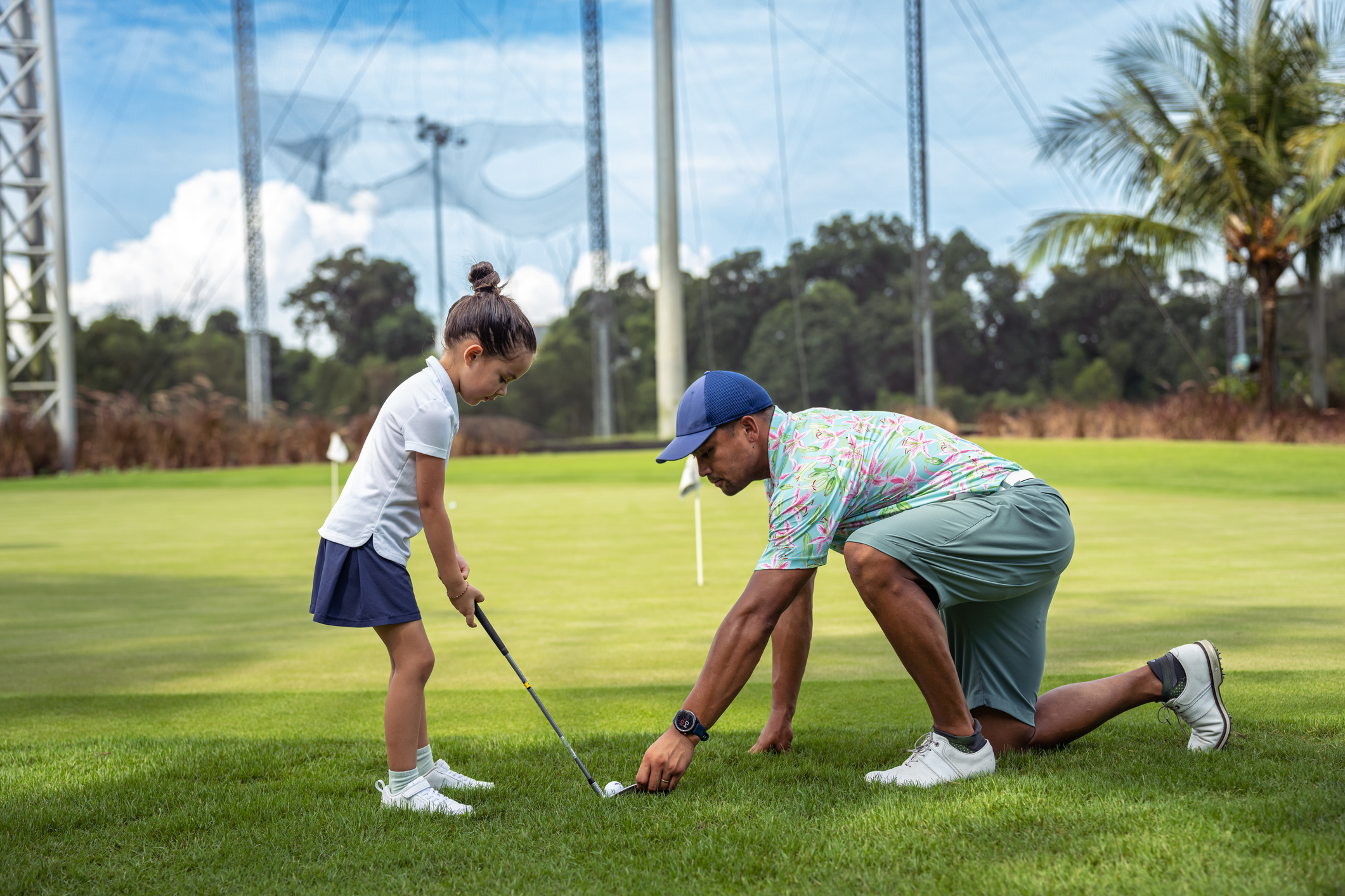 Un père s'agenouille sur le terrain de golf, repositionnant une balle de golf pendant qu'il enseigne à sa fille de maternelle comment putter.