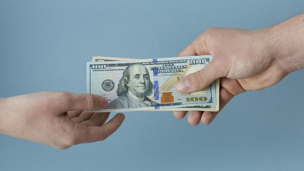 A man's hand holds out several hundred-dollar bills to the hand of a younger person.