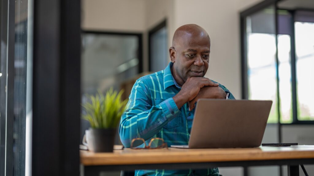A man appears to be thinking while sitting at his desk in front of his laptop.