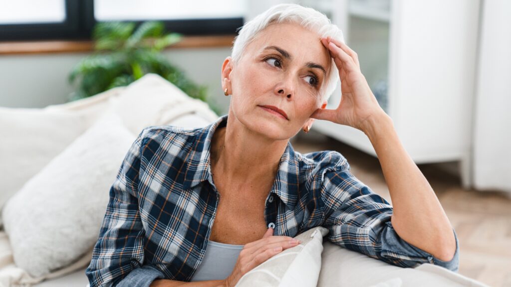 An older woman looks stressed as she sits on her sofa and looks into the distance.