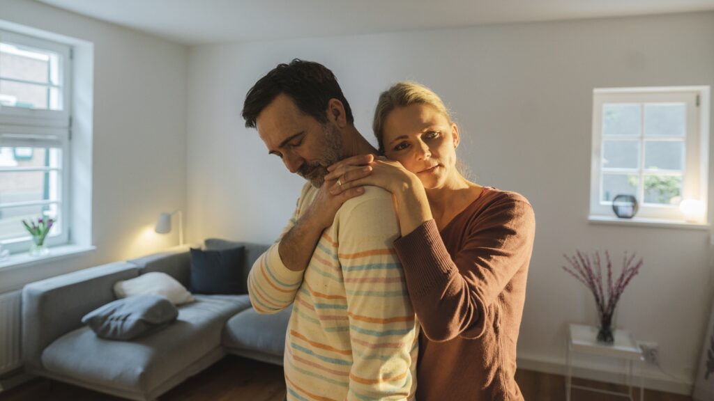 A man and woman embrace in their living room, looking concerned.