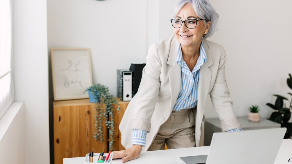 An older woman smiles as she looks off to the side while standing at her desk in an office.