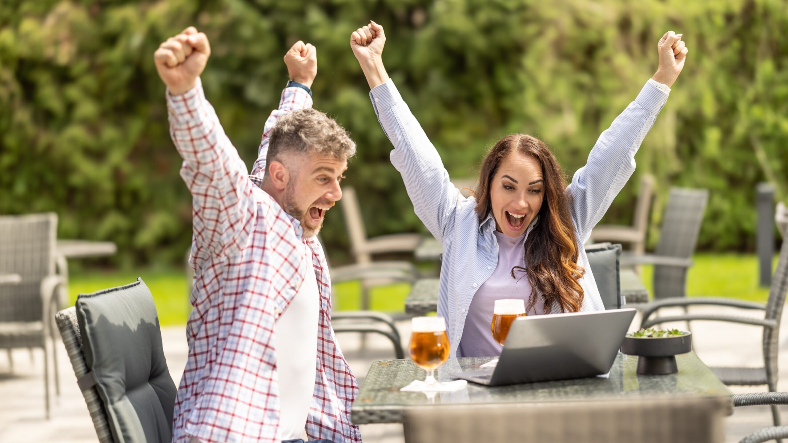 A man and woman celebrate a win as they sit at an outdoor cafe looking at a laptop.