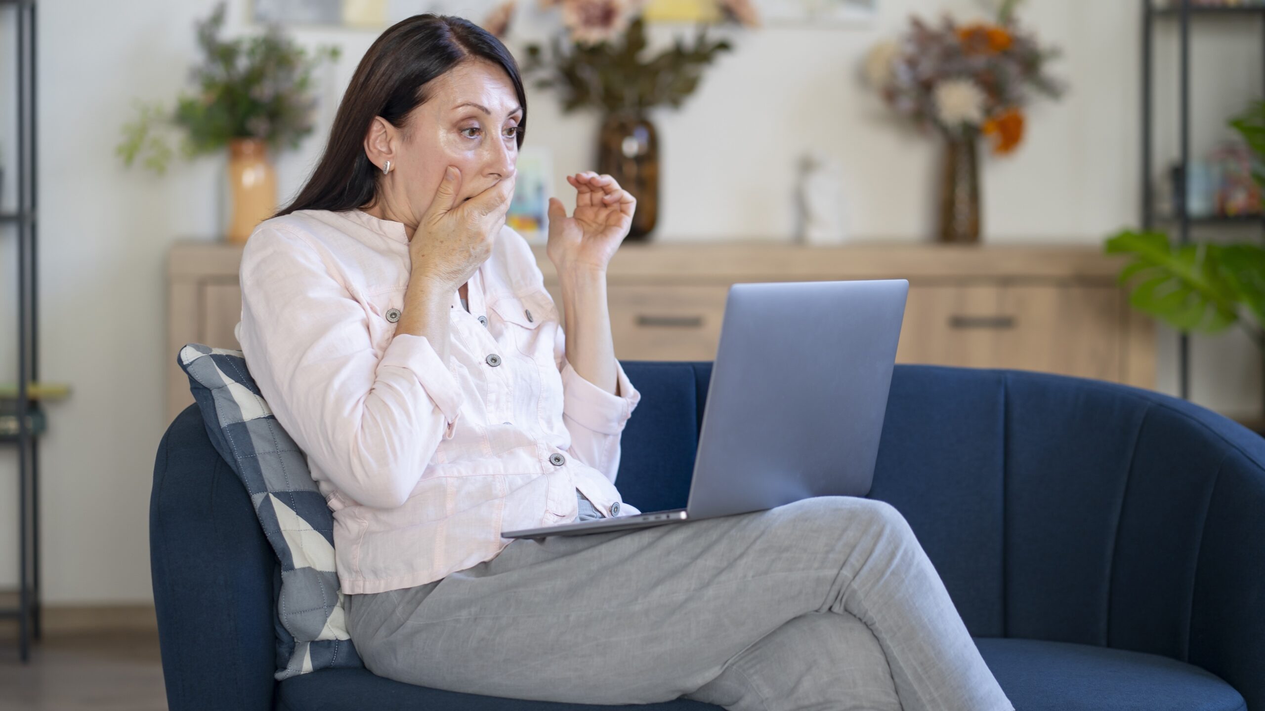An older woman covers her mouth in shock as she looks at her laptop on her sofa.