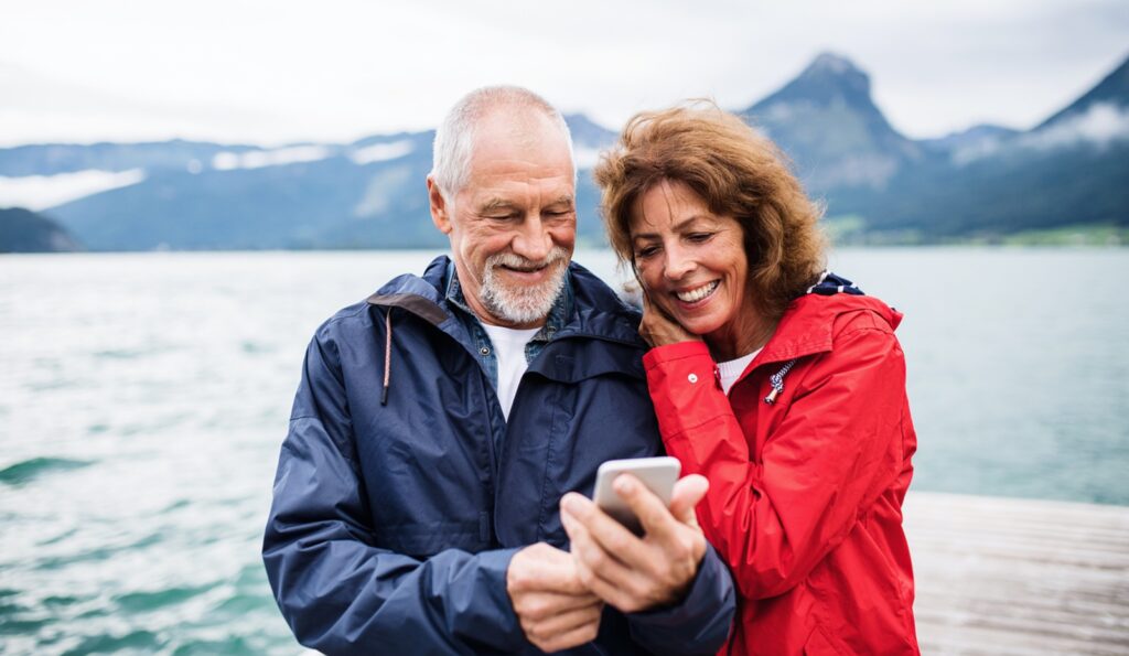 A mature couple on vacation in a beautiful, mountain location on the water look at a cell phone.