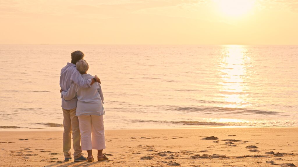 An older couple stand arm in arm on a beach and watch the sunset.
