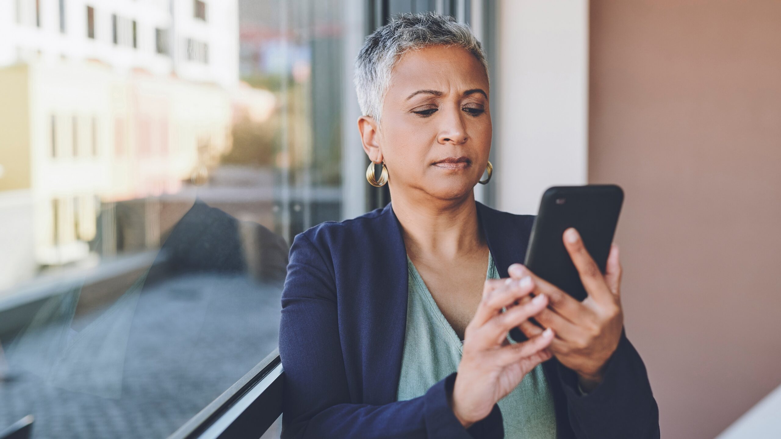 A businesswoman looks skeptical as she looks at her phone while standing near her office window.