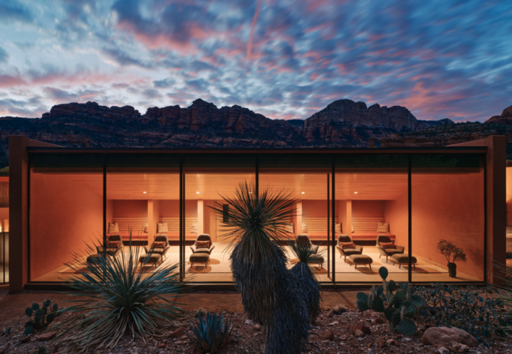 The image shows a relaxation room at Mii Amo spa, lit up at sunset. In the foreground are cacti and in the background, mountains and a multi-colored sunset.