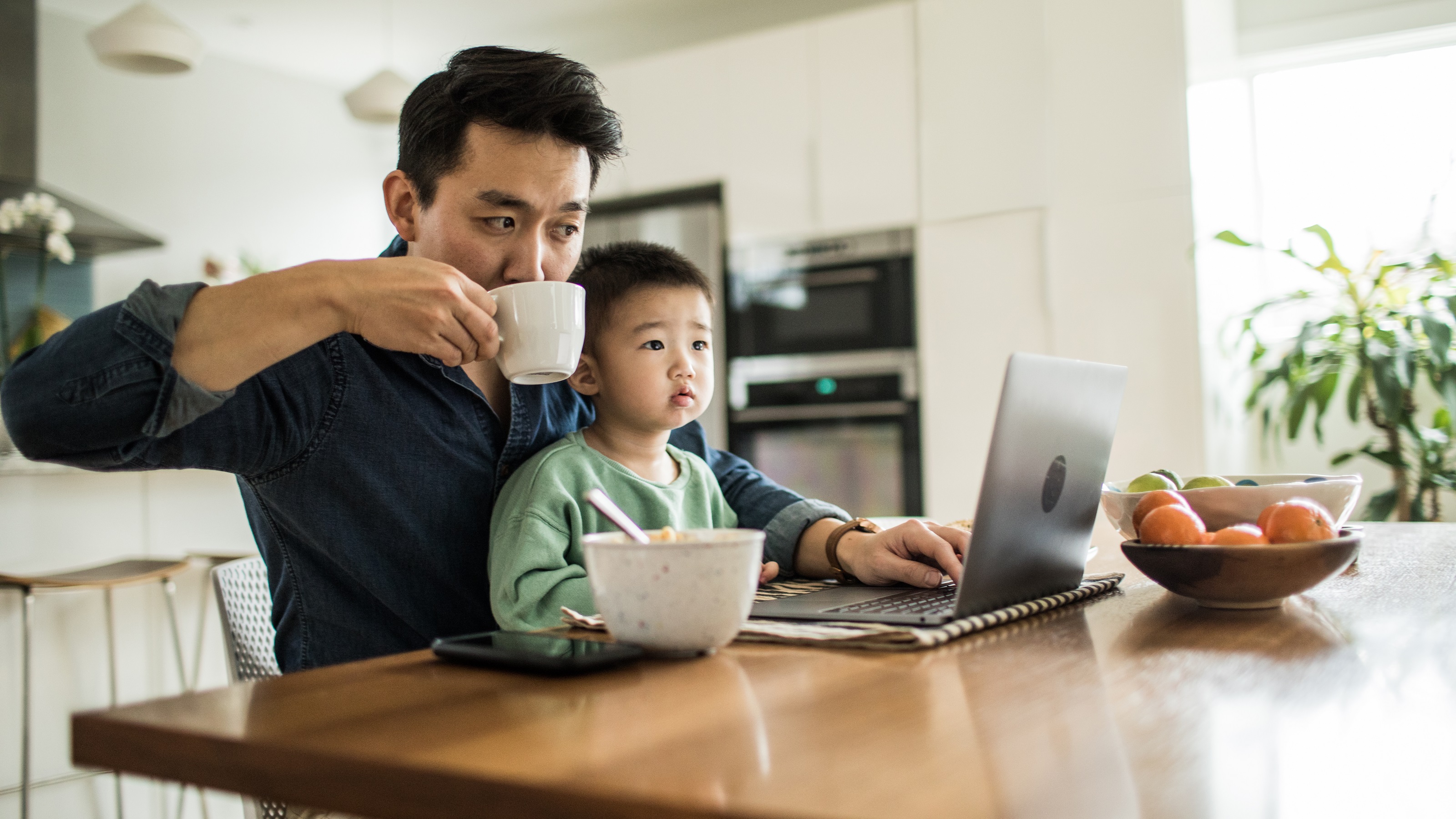 Un père travaille sur son ordinateur portable à la table de la cuisine pendant que son jeune fils est assis sur ses genoux