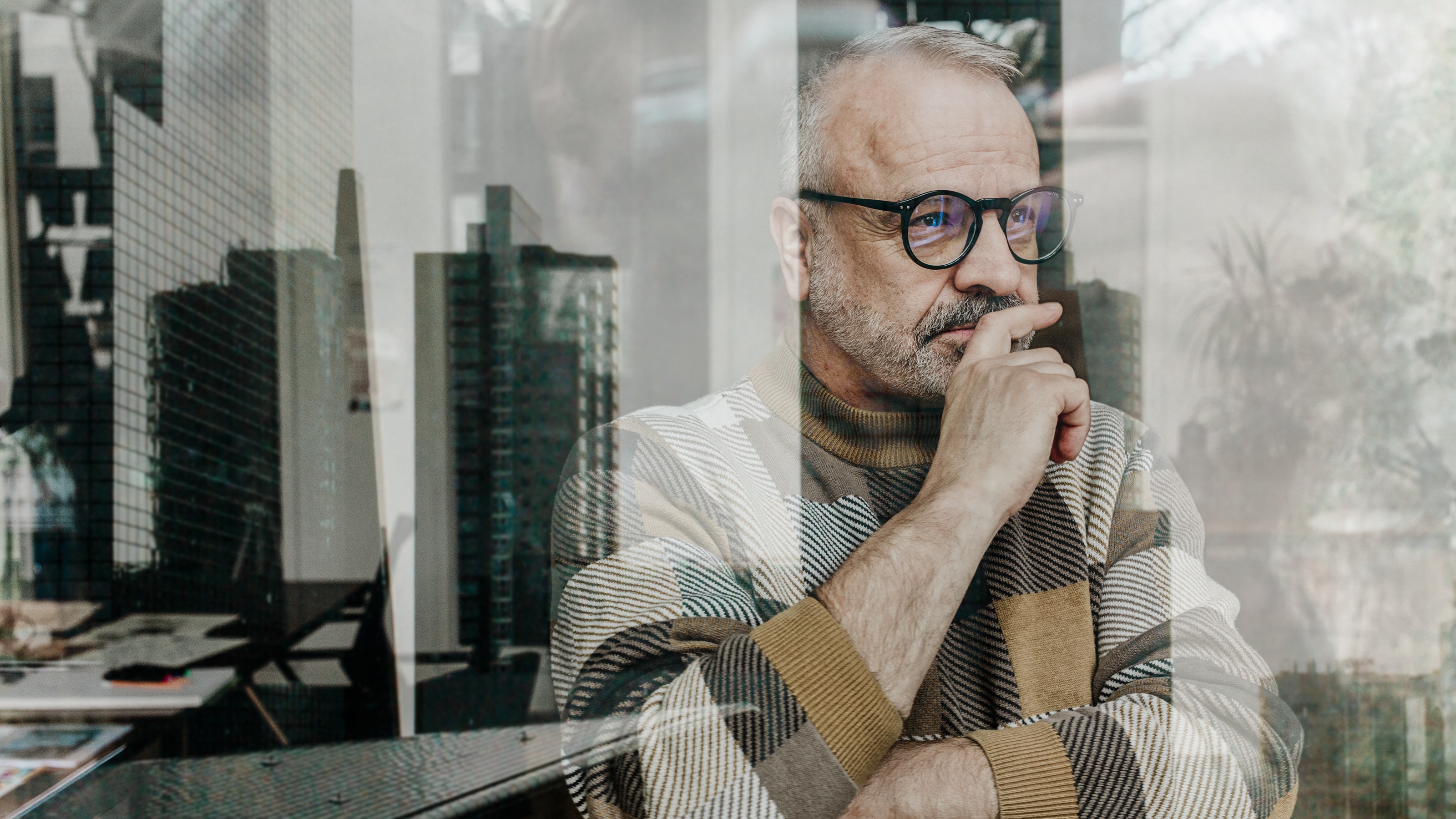 Un homme plus âgé a l’air pensif alors qu’il regarde par la fenêtre de son bureau, une ligne d’horizon se reflétant dans la vitre.