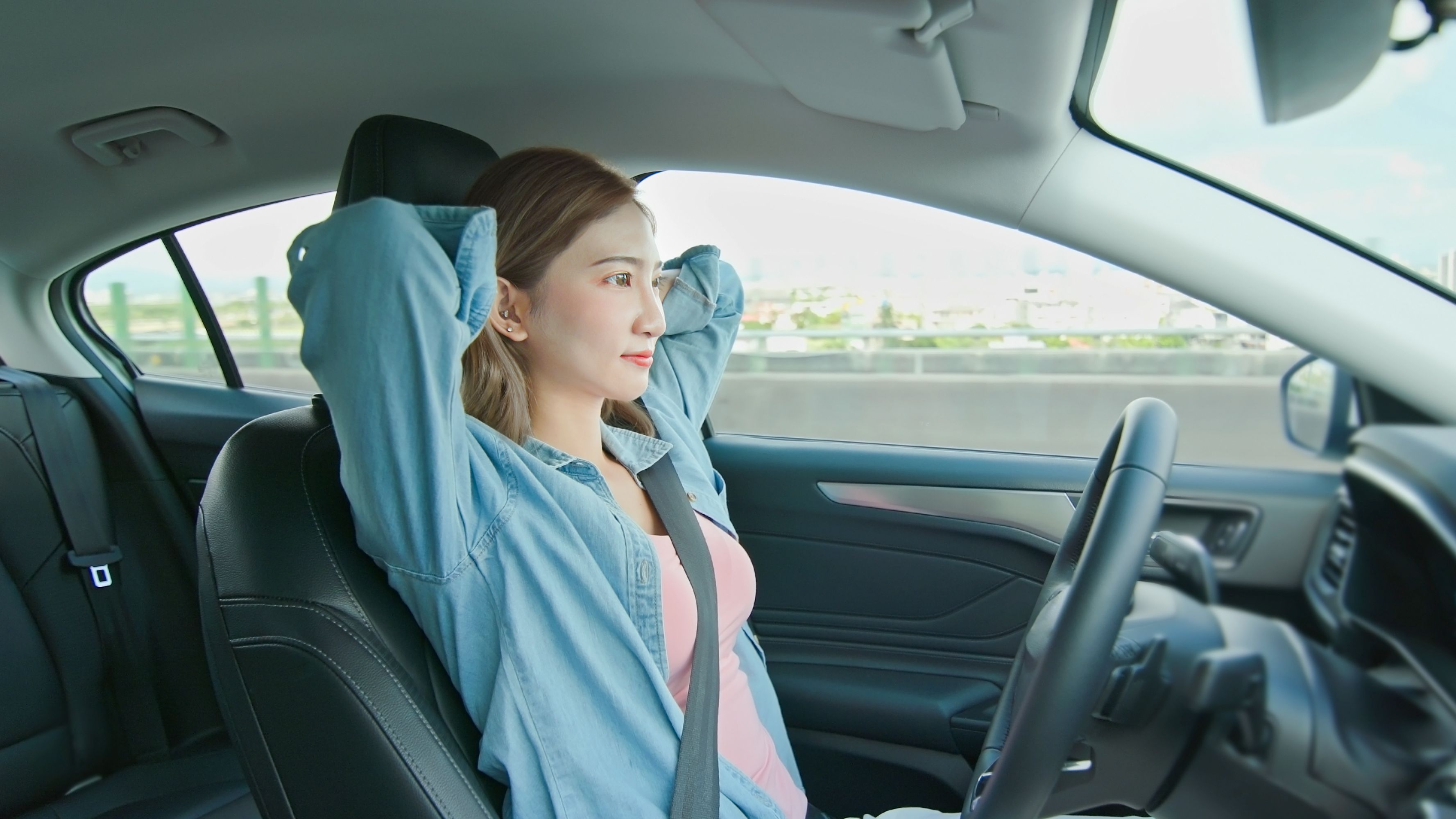 Une femme est assise sur le siège conducteur, les mains derrière la tête, car elle utilise la fonction de conduite autonome de sa voiture.