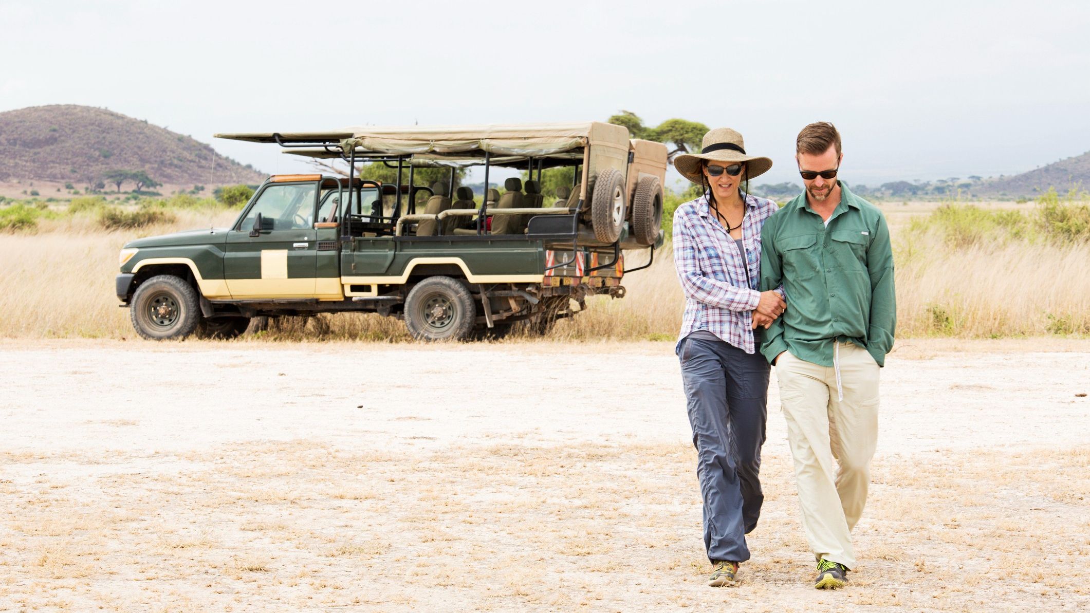 Un couple d’âge moyen portant des lunettes de soleil s’éloigne d’une grande jeep de safari.