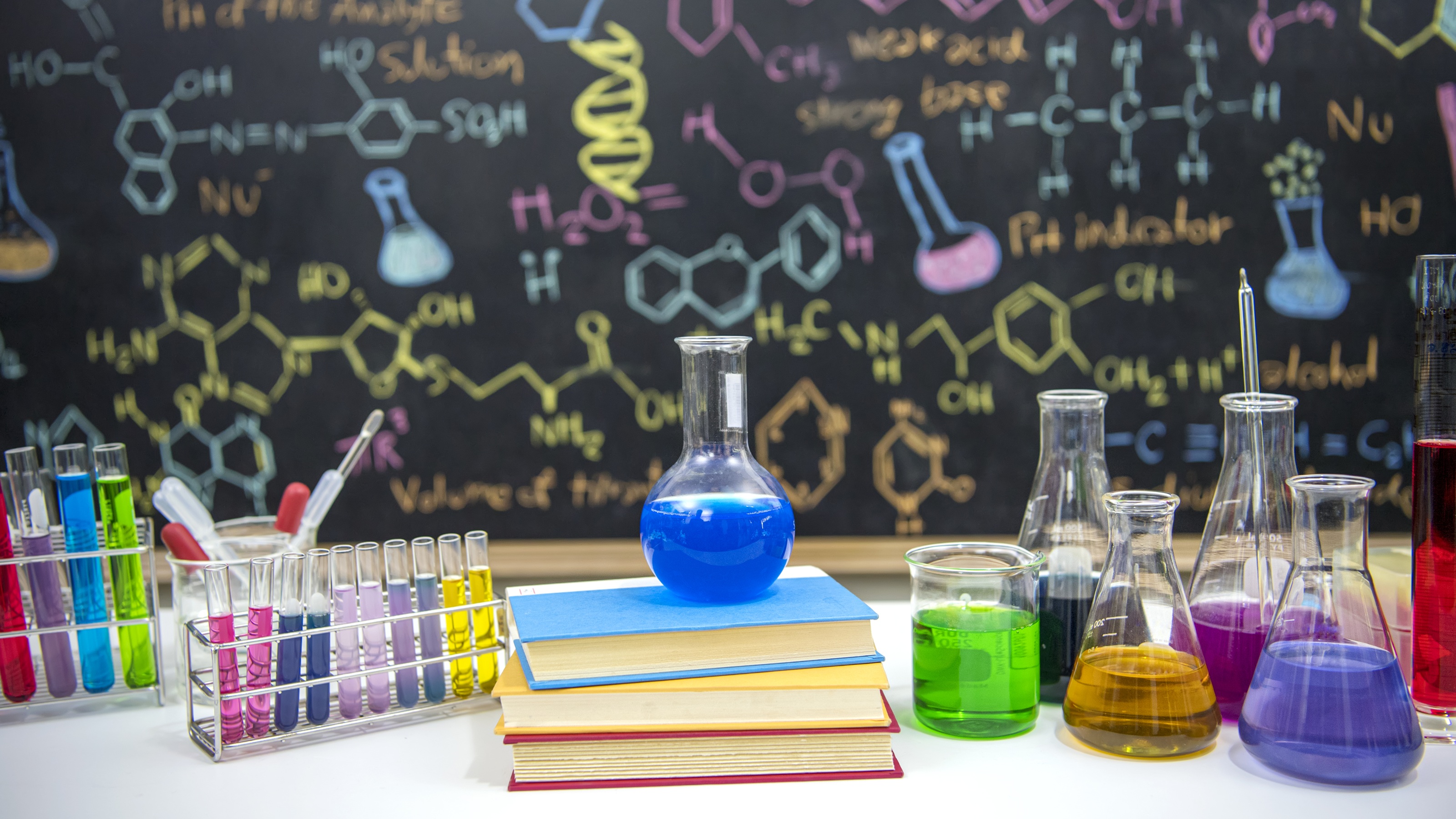 Bureau de classe avec livres et instruments scientifiques devant un tableau avec une formule dessinée à la craie colorée.