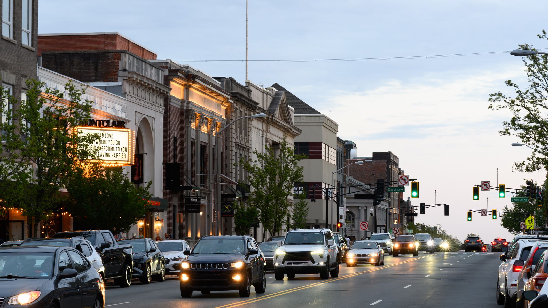 Les voitures circulent sur la route à Montclair, dans le New Jersey, traversant un centre-ville bordé de bâtiments historiques en brique et de devantures de magasins locaux, dont un théâtre avec un chapiteau illuminé.