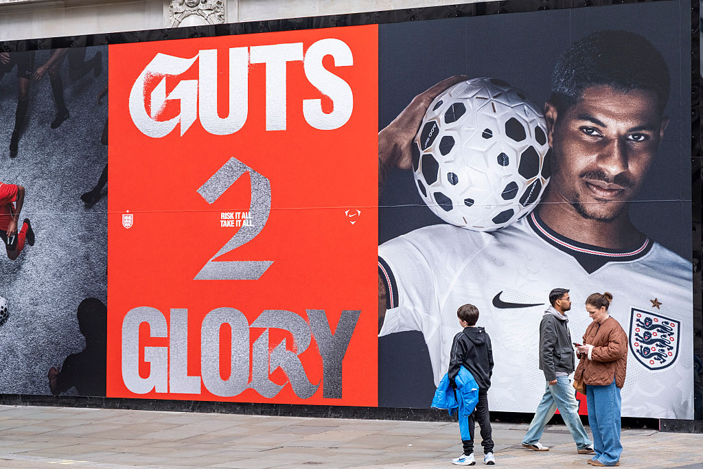 Des gens debout à côté d’une fresque murale de la Coupe du monde qui dit : "Guts 2 Gloire" et il y a un joueur de football qui porte un maillot Nike