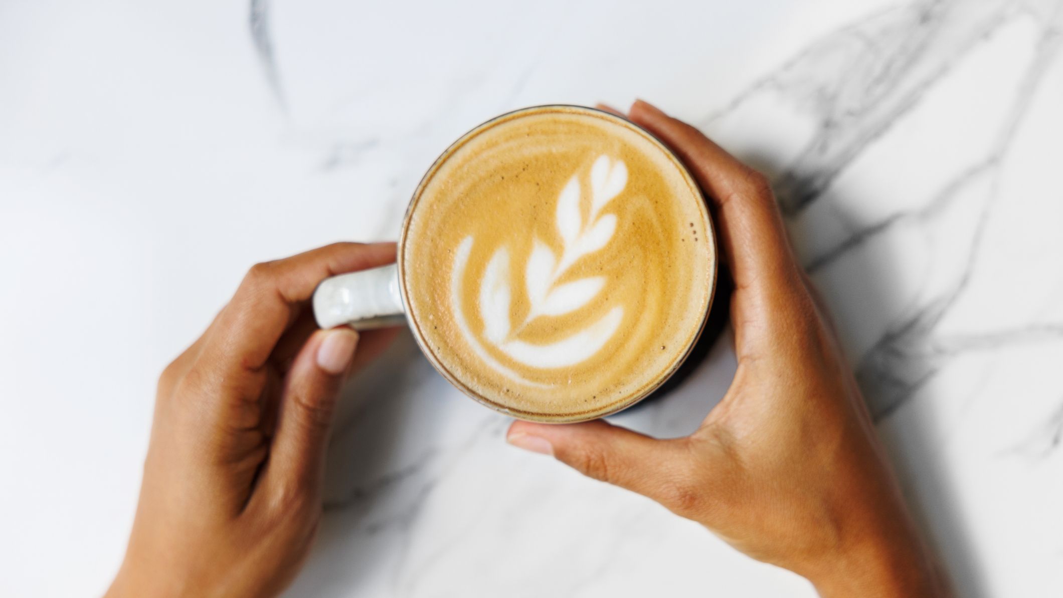 Mains tenant doucement une tasse de café fraîchement infusée avec un latte art complexe sur une surface en marbre.