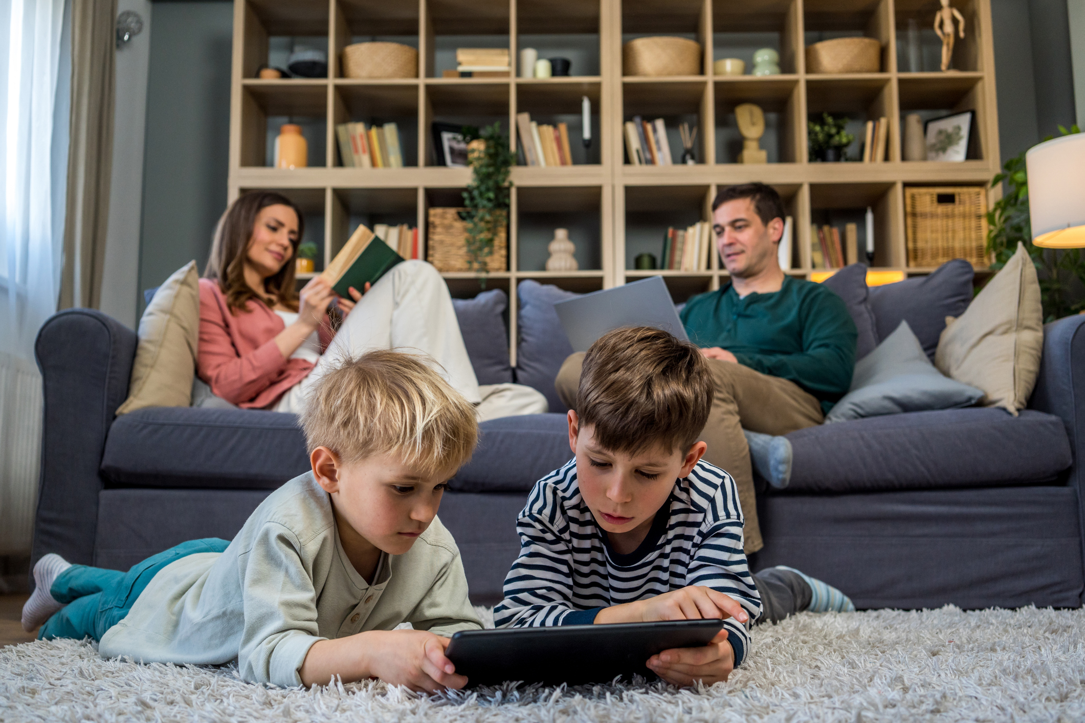 Deux fils jouent à des jeux sur une tablette posée au sol pendant que leurs parents se détendent sur le canapé avec un ordinateur portable et un livre, profitant du temps passé en famille à la maison.