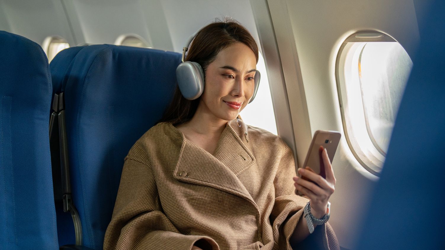 On voit une femme chic se détendre au siège près de la fenêtre d'un vol, profitant de son temps avec des écouteurs sans fil et un smartphone