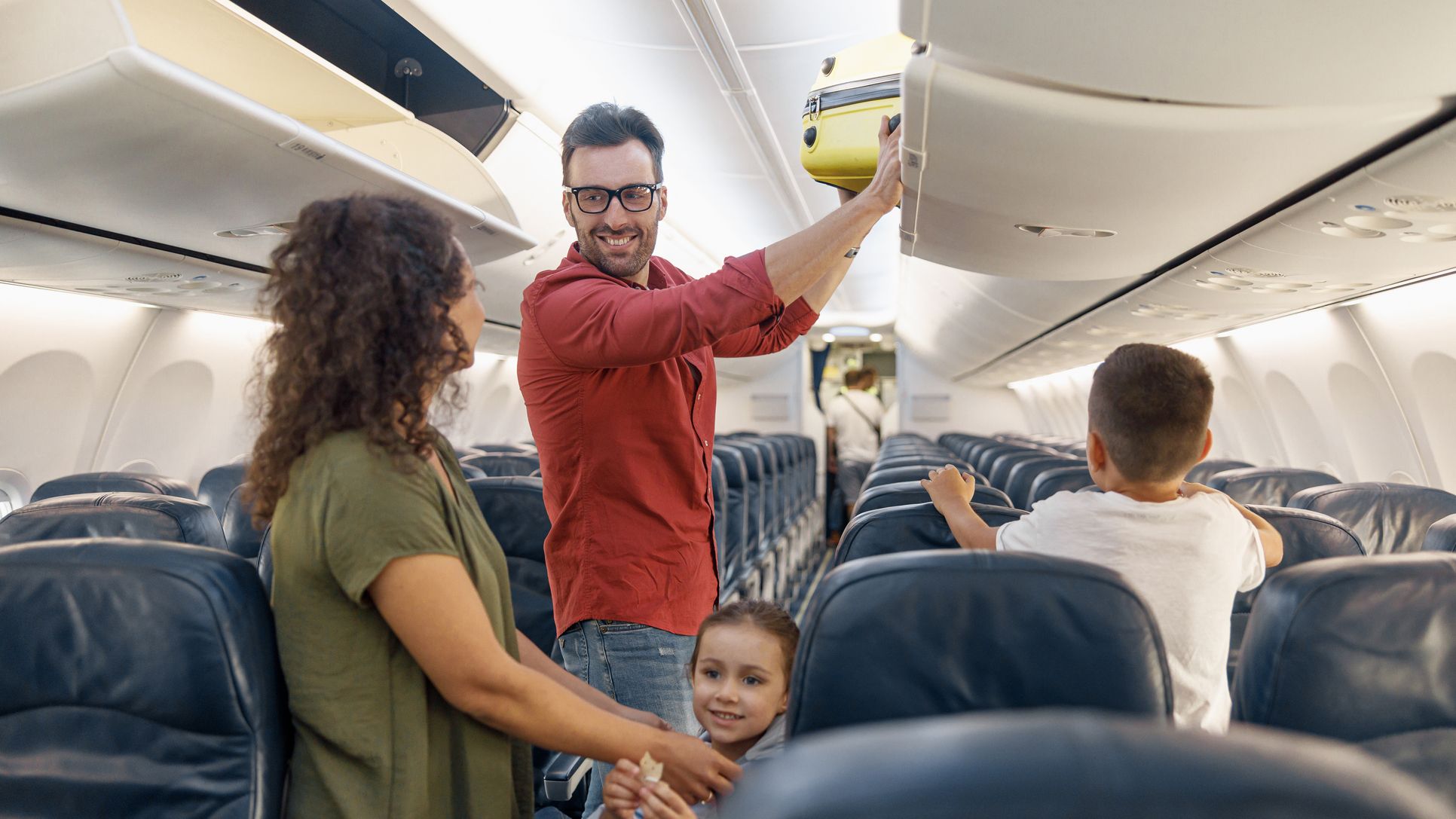 Homme joyeux regardant sa femme avec un sourire et mettant ses bagages à main dans le compartiment tout en voyageant avec sa famille en avion