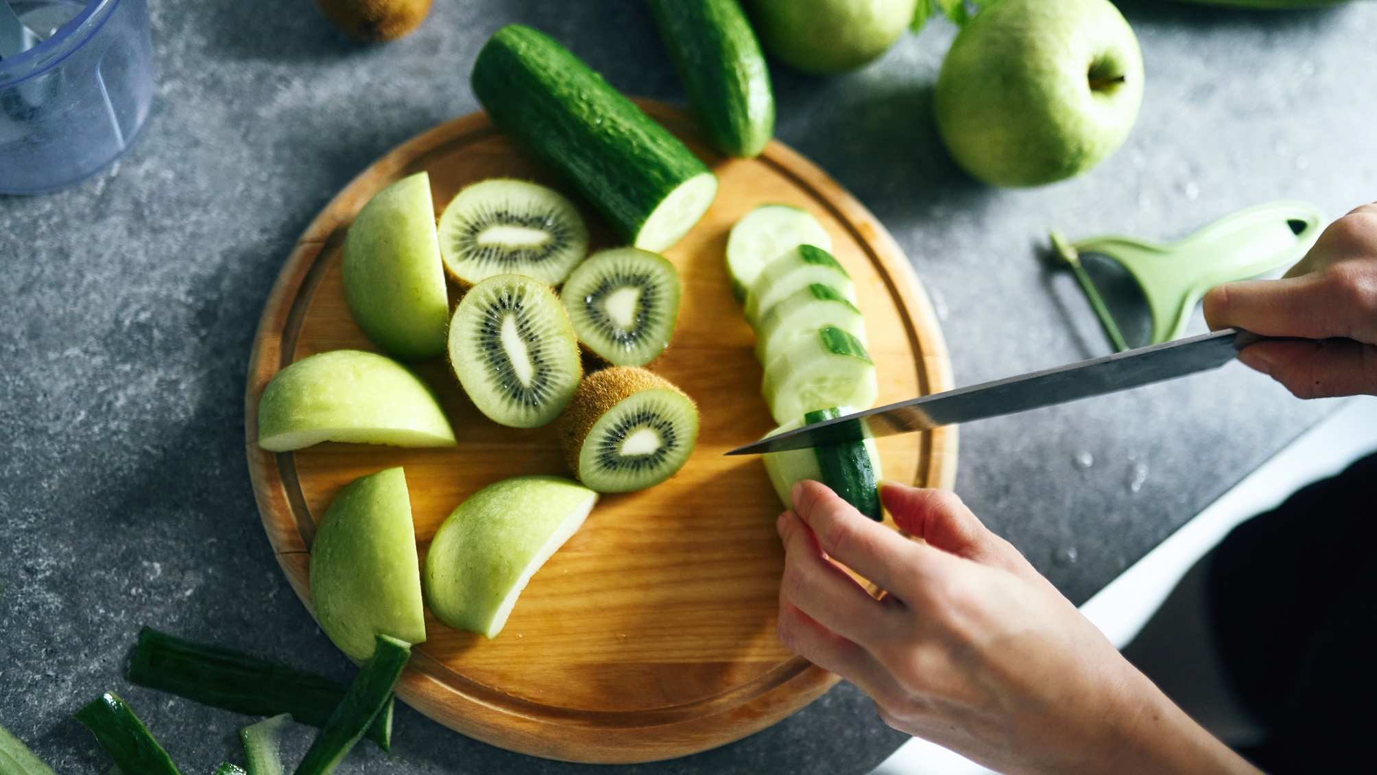 Mains coupant un concombre sur une planche à découper avec du kiwi et de la pomme verte.