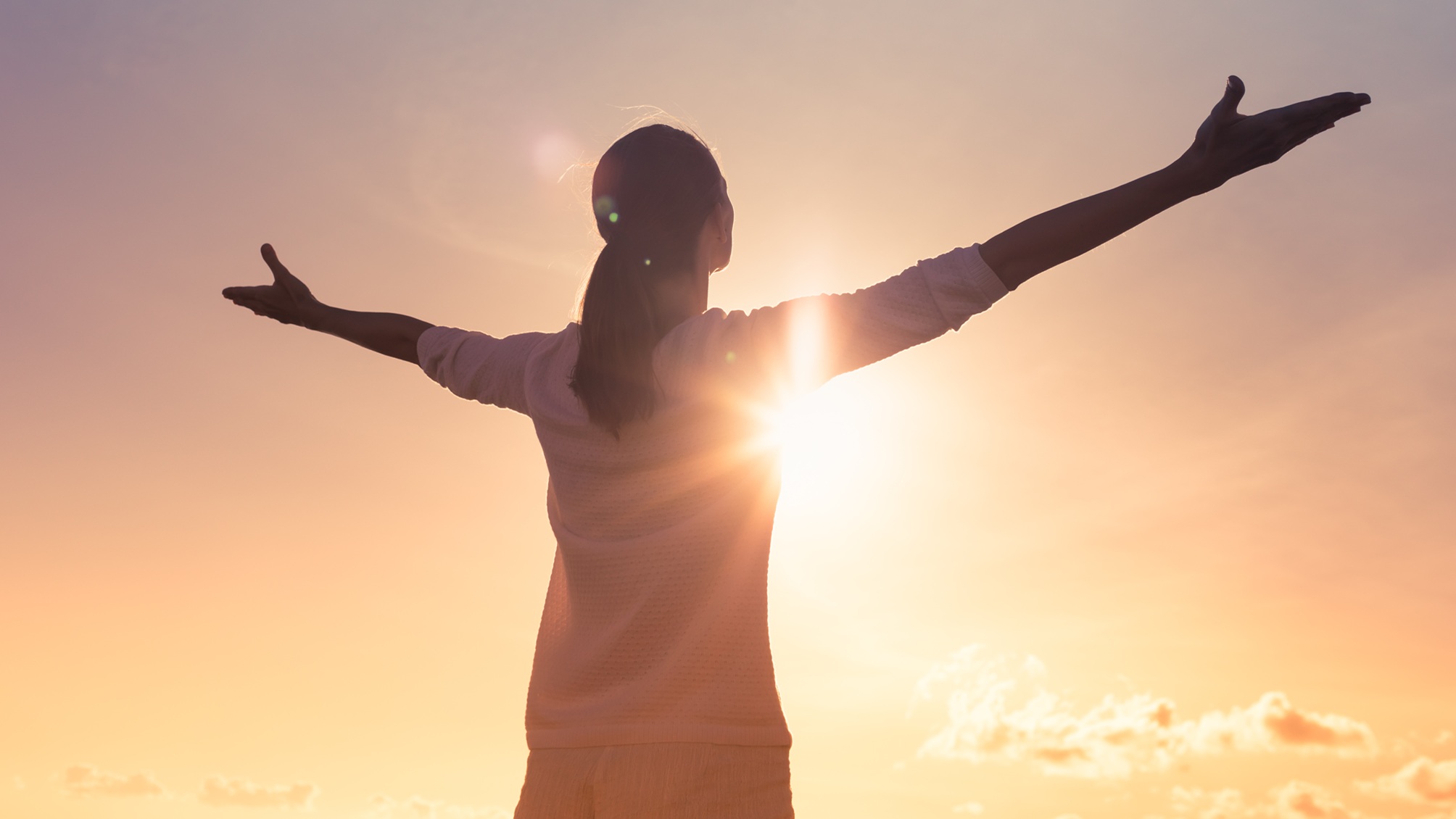 Une femme fait face à un coucher de soleil et écarte les bras de manière détendue.