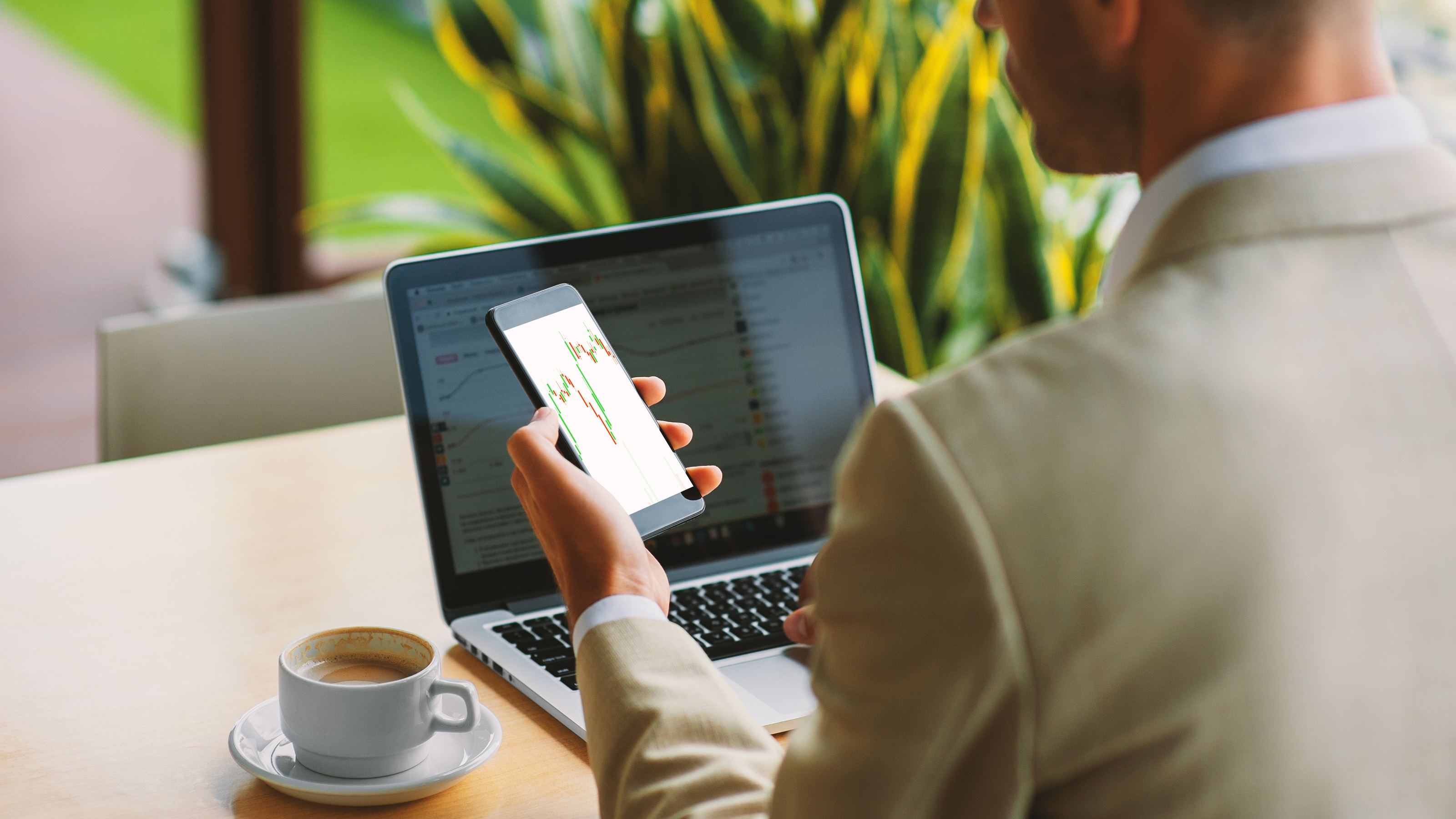 Un homme regarde un graphique de négociation d'actions sur son téléphone alors qu'il est assis à une table de café avec son ordinateur portable ouvert.