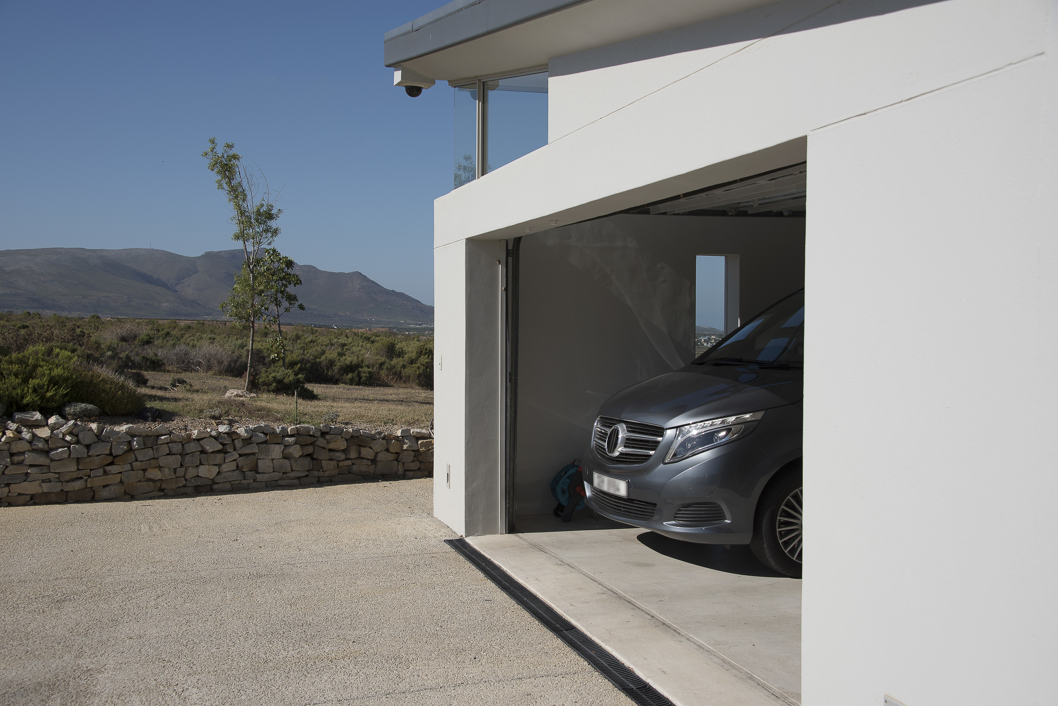 Un garage dans une maison moderne abrite une voiture de luxe. Il y a des montagnes au loin.