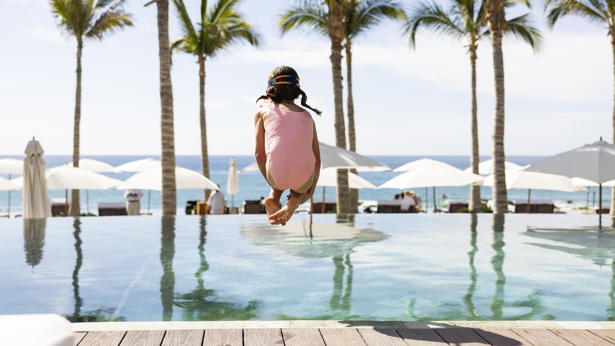 Une jeune fille saute dans une piscine, une plage et des palmiers en arrière-plan.