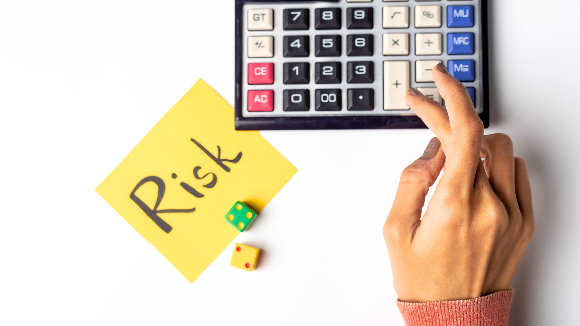 Une main utilisant une calculatrice à côté d'un pense-bête et d'un dé. La note a "Risque" écrit dessus.