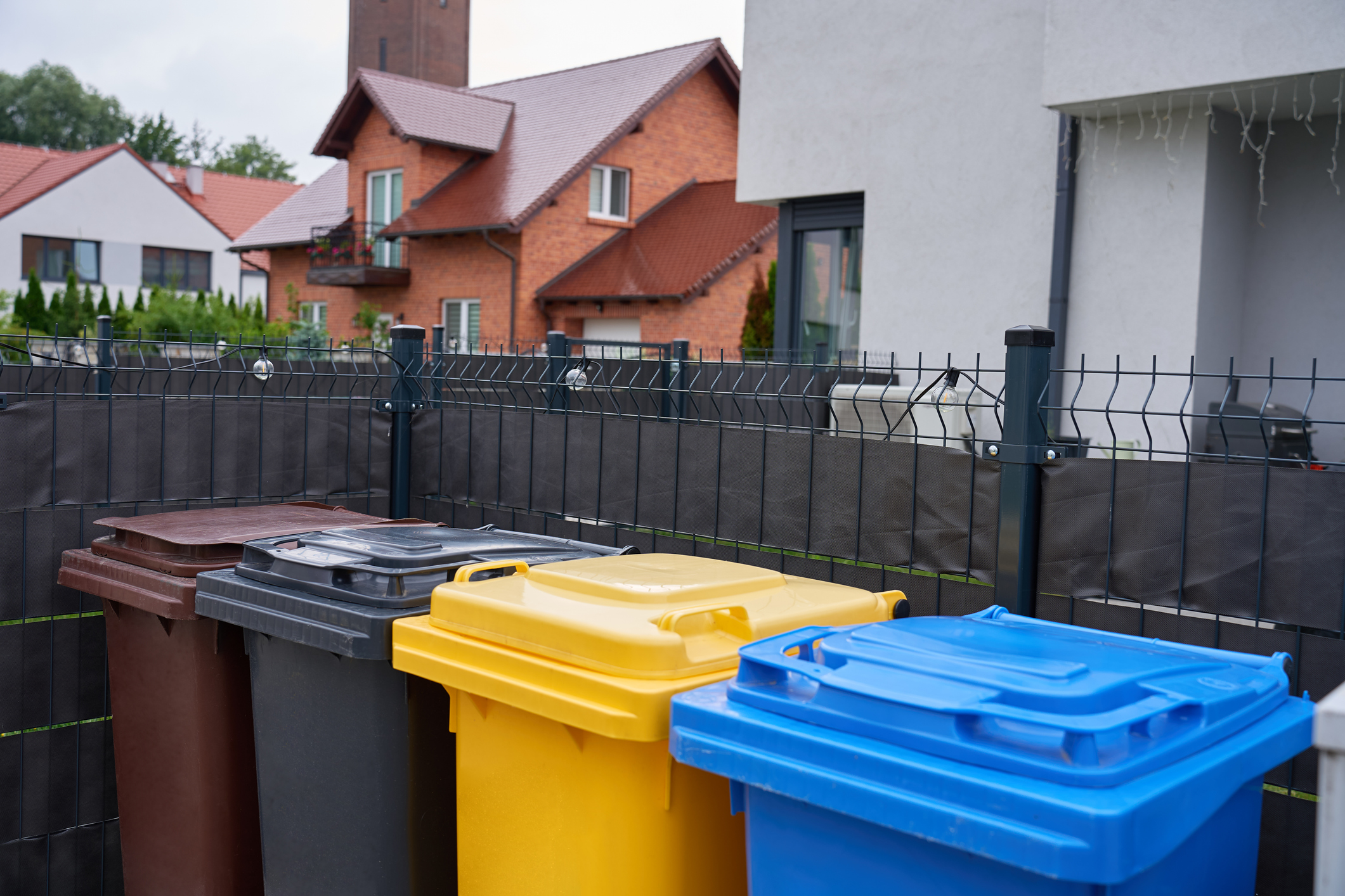 Des bacs de recyclage colorés se trouvent derrière une clôture dans un quartier résidentiel à proximité de maisons modernes.