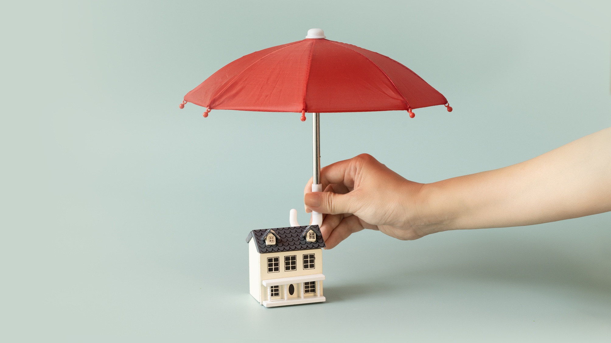Une main tient un parapluie rouge au-dessus d'une maison de jouets.