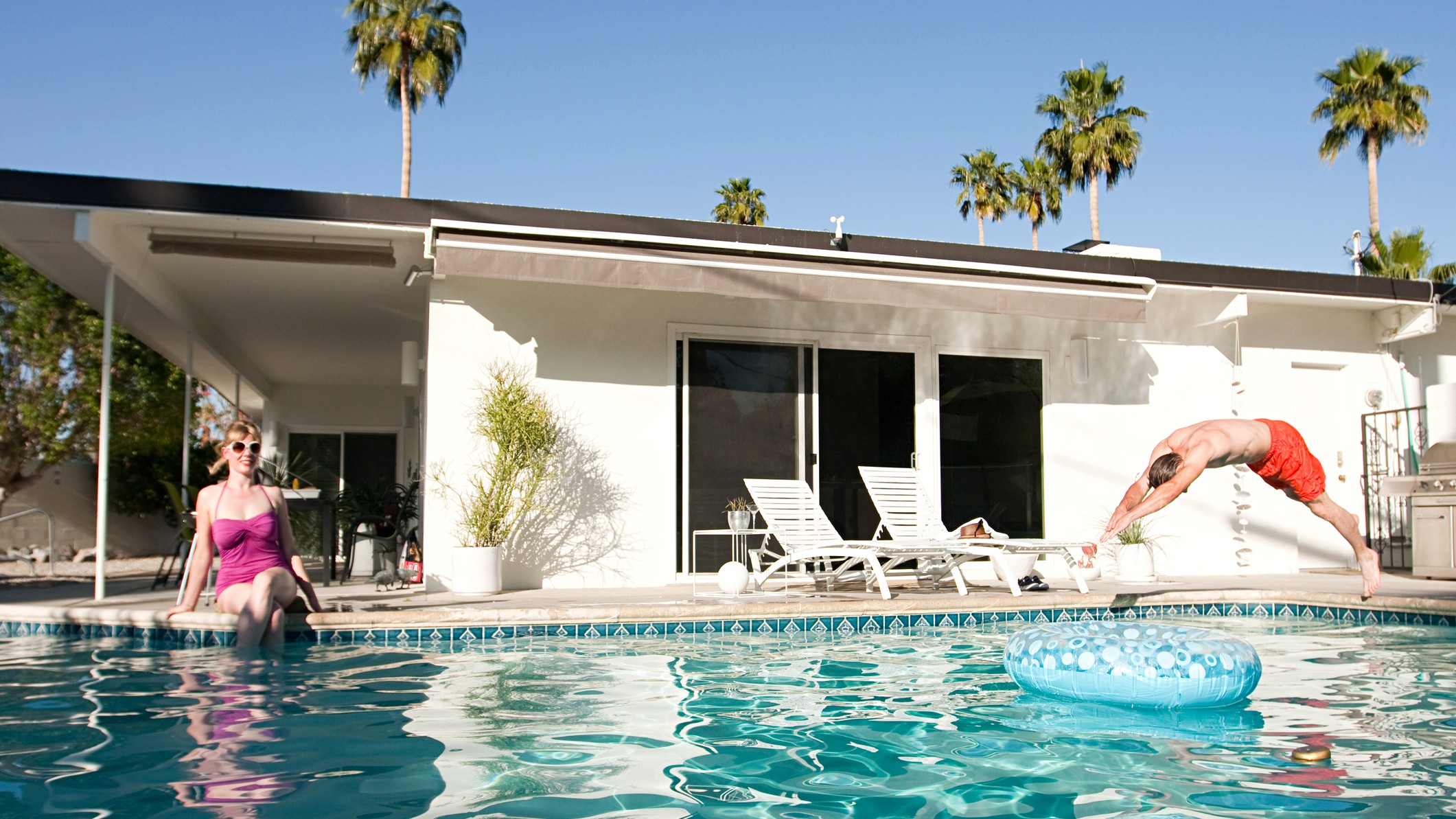 Une maison moderne et une piscine à Palm Springs, en Californie. Deux adultes nagent, vêtus de maillots de bain aux couleurs vives pour une ambiance rétro.